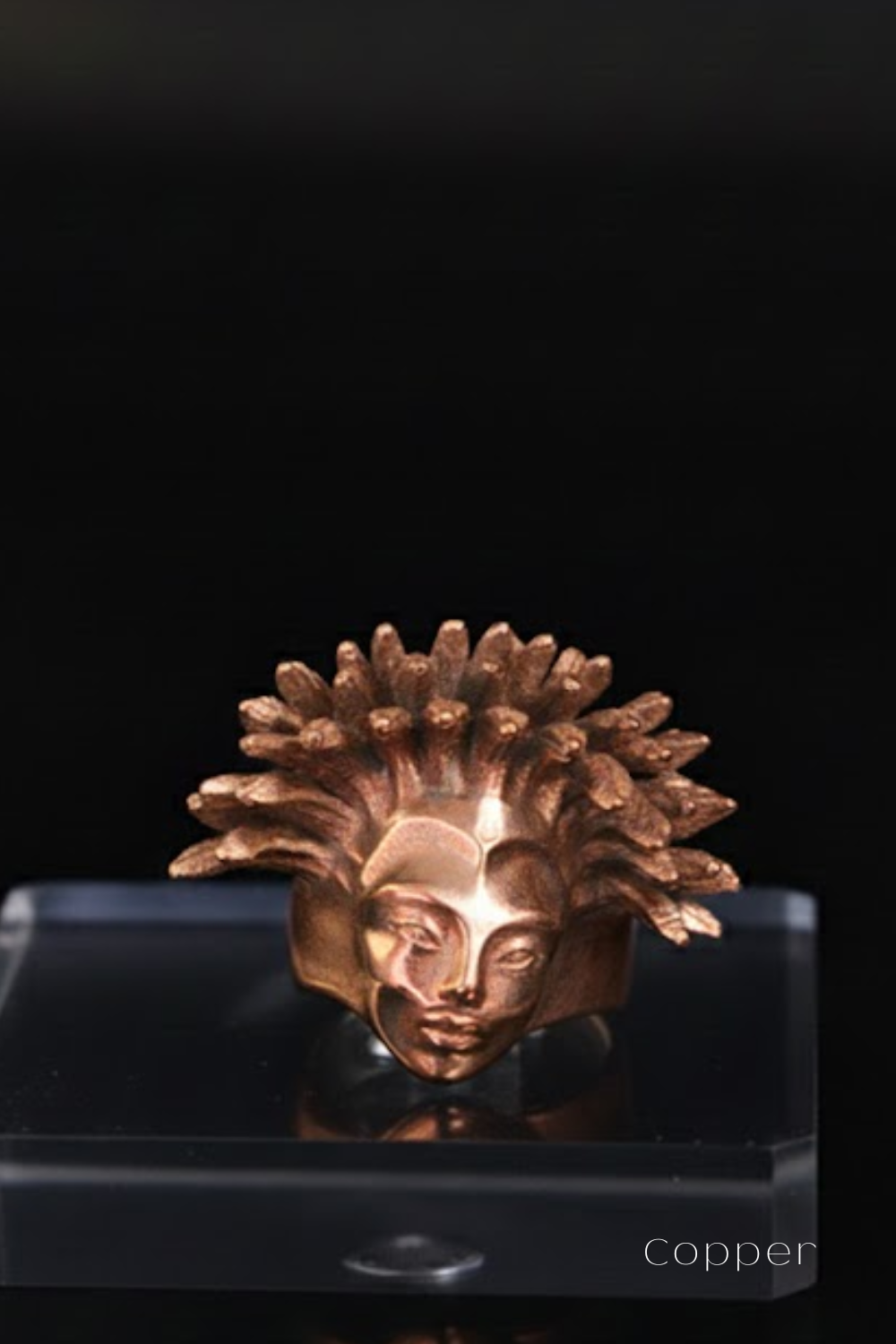 A copper-colored ring with an intricate, stylized face design is displayed on a clear stand against a black background.