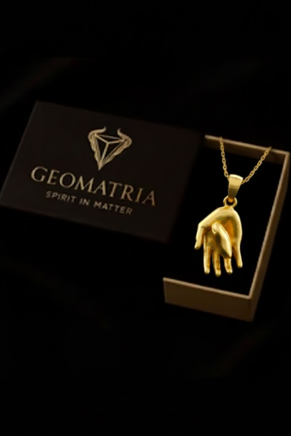 A gold-colored pendant necklace is displayed in an open box with the text "Geomatria" and "SPIRIT IN MATTERS" printed on it.
