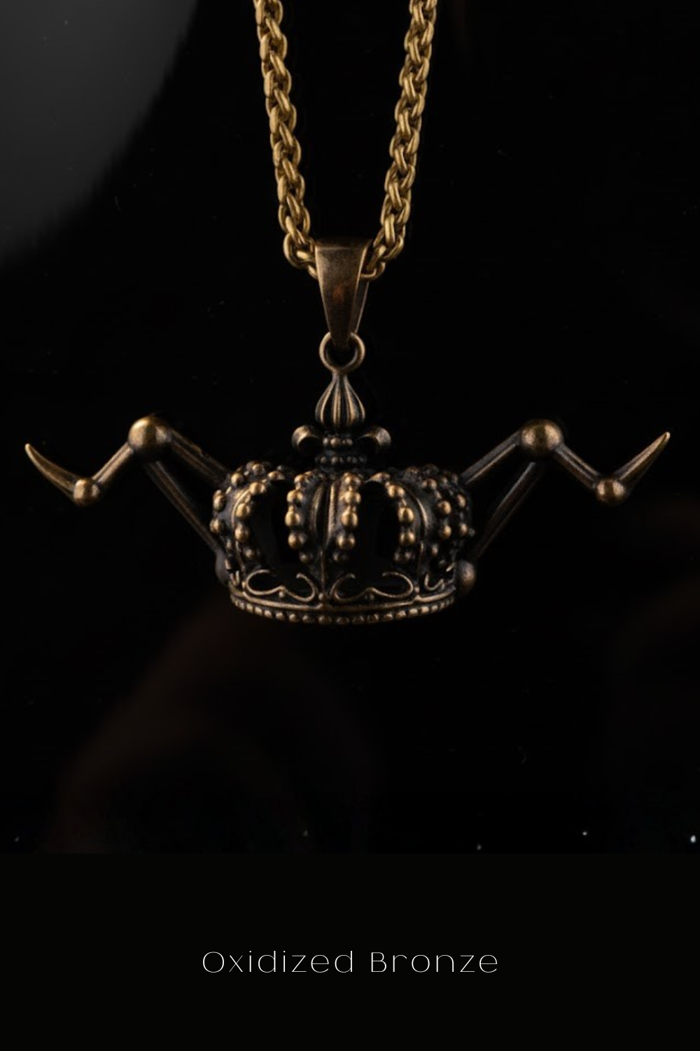 An oxidized bronze pendant in the shape of a crown, suspended from a chain against a black background.