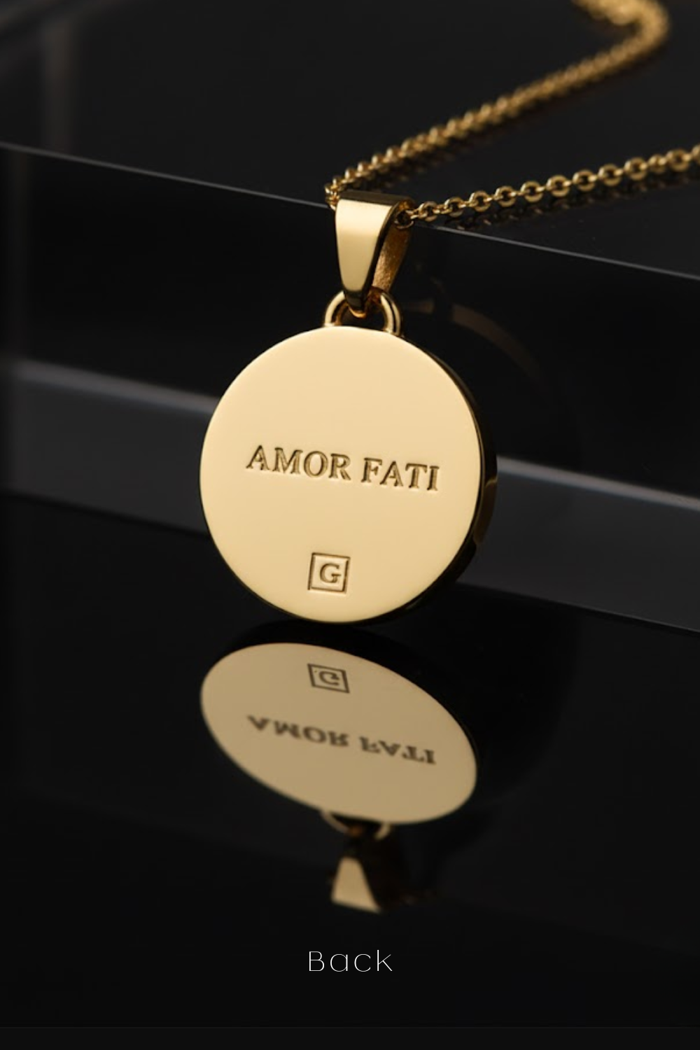 a gold-colored pendant necklace with the text "Amor Fati" engraved on it, displayed on a black surface.