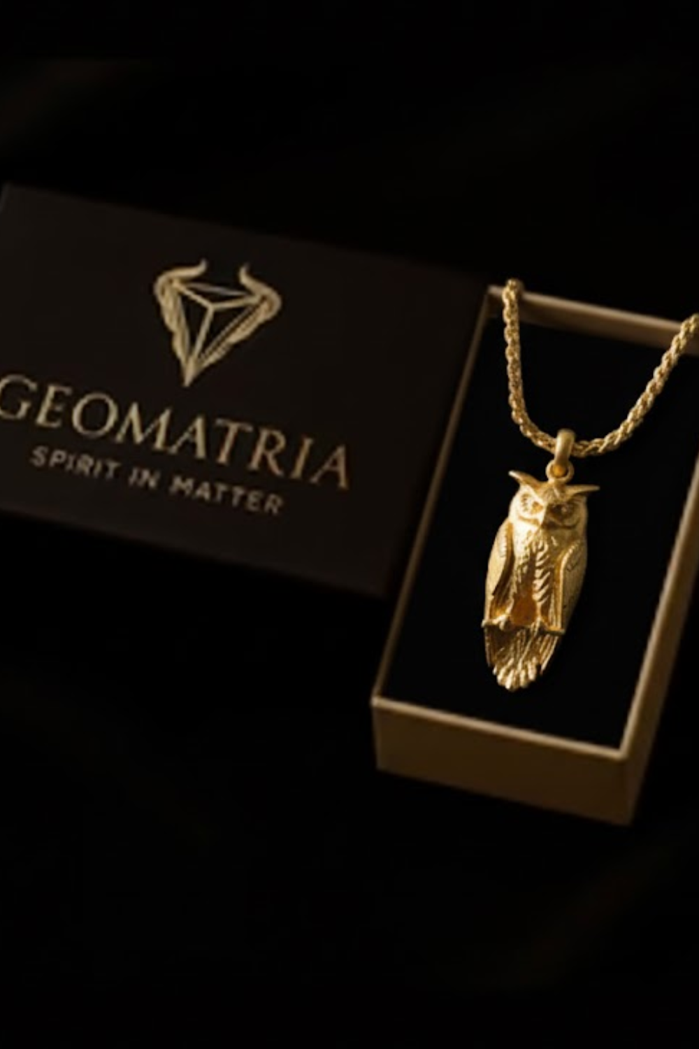 A gold-colored owl pendant is displayed in a gold-colored box with the text "Geomatria" and "Spirit in Matter" printed on it.