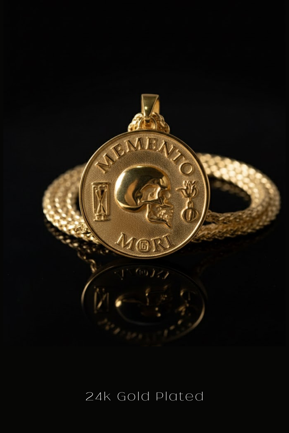 A 24k gold plated pendant with a skull design, accompanied by the Latin motto "MEMENTO MORI" (Memento mori) and the word "MORI" (motion of death). The pendant is displayed on a chain against a black background.