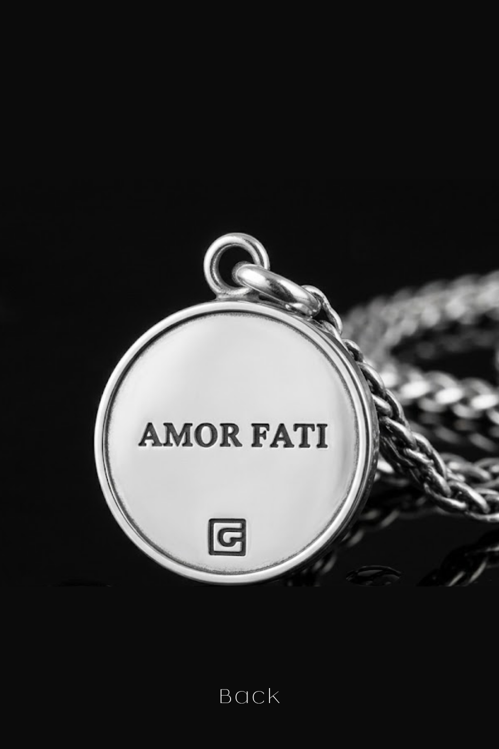 a silver-colored pendant with the text "Amor Fati" engraved on it, set against a black background.