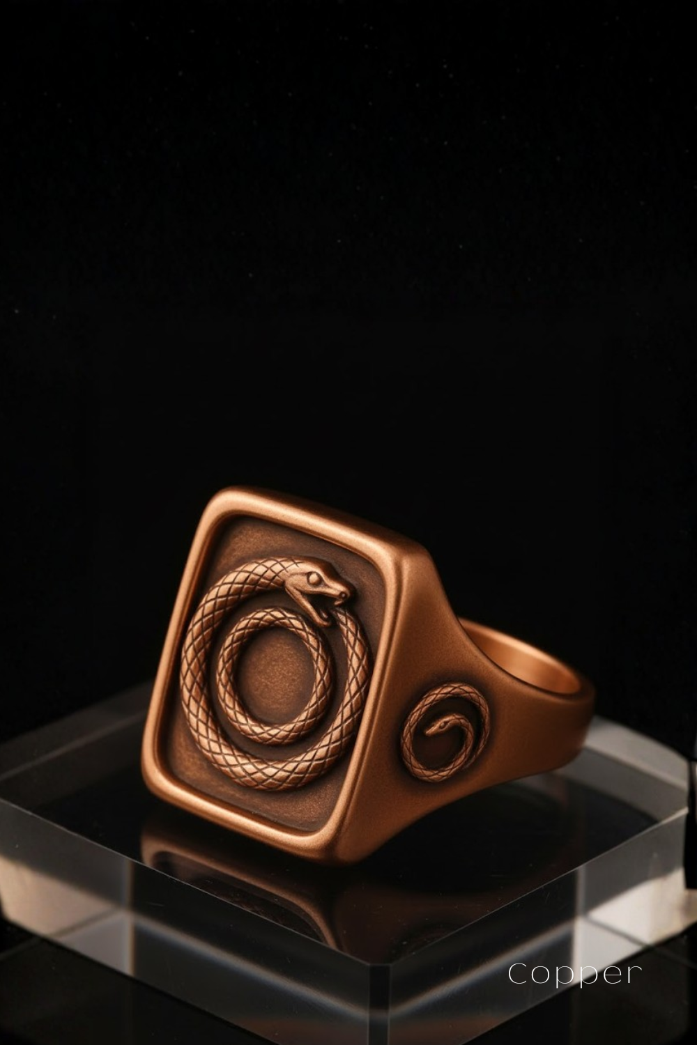 A copper-colored ring with a square design featuring a snake motif, displayed on a clear glass stand against a black background.