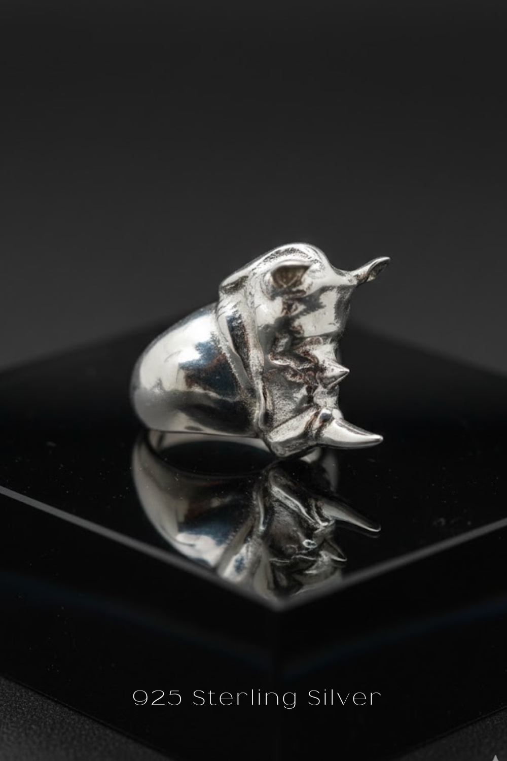 A silver ring with a unique design sits on a reflective surface against a dark background.